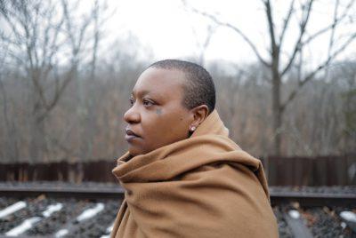 Musician Meshell Ndegeocello, a Black woman with shortly cropped natural hair and a star tatttooed near her right eye, stands outside near railroad tracks in the late fall or winter, wrapped in a camel-colored coat or large scarf. She looks towards the right of the frame, and some of the wooden tithes are covered in melting snow behind her.