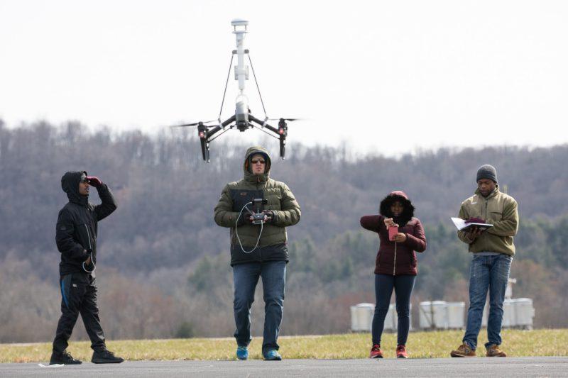Undergraduates from Bennett, 豪斯, and Hampden Sydney stand on an airstrip at Virginia Tech’s Kentland Farm. David Schmale is manning a drone. One student is writing in a notebook, another is recording a video on her phone, and another is observing the drone and protecting his eyes with his hands. Courtesy of Peter Means.